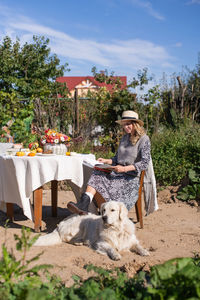 A woman in a hat is sitting on a chair near the table, a labrador retriever is lying next to her