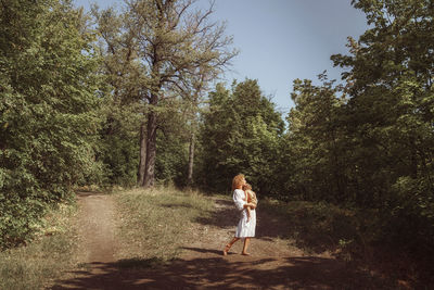 Woman holding a baby son standing by plants in forest against sky