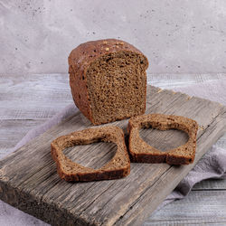 Loaf of rye bread and two slices with carved holes of heart shape on old wooden board on grey. 