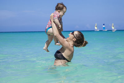 Side view of cheerful woman carrying toddler daughter in sea against blue sky