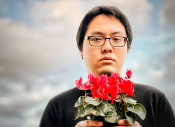 Portrait of young man holding red flowering cyclamen plant against cloudy blue sky.