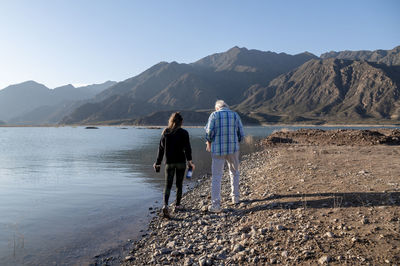 Senior man walking with his granddaughter along the lake shore with mountains.