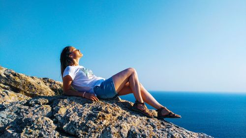 Woman sitting on rock by sea against clear blue sky