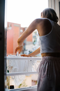 Back view of unrecognizable female in domestic outfit standing near window and attaching wet clothes with clothespins while doing household chores at weekend