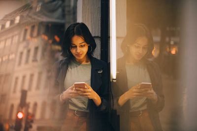 Young woman texting through phone while standing by glass window