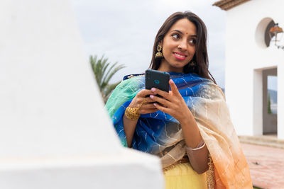 From below satisfied indian woman in traditional outfit texting via mobile phone while standing in garden