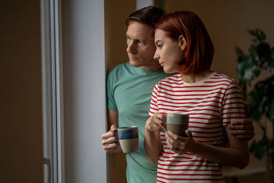 Man and woman with mugs of coffee look out window. reluctance to leave house, day off, weekend