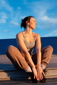 Full body of female in sportswear looking into distance while sitting on wooden stair during training on sunny street in city