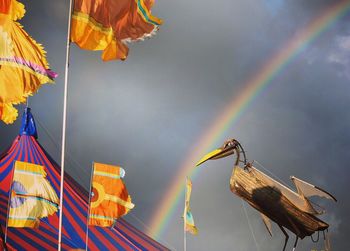 Low angle view of artificial bird by flags against cloudy sky with rainbow
