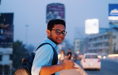 Side view portrait of smiling of young man with backpack standing on street at dusk