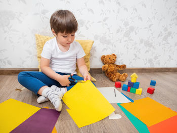 Toddler boy learns to cut colored paper with scissors. kid sits with toy blocks and teddy bear. 