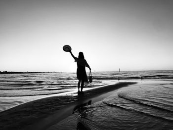 Silhouette woman holding hat standing on beach against clear sky