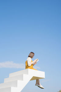 Low angle view of woman sitting on steps while reading book against sky