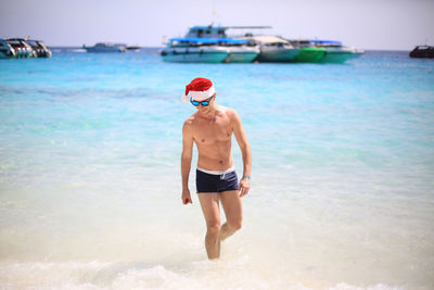 Shirtless man wearing santa hat while wading in sea against sky