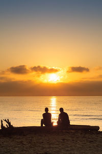 Silhouette people sitting on beach against sky during sunset