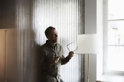 Mature businessman holding eyeglasses while talking on smart phone through headphones standing against wall at creative