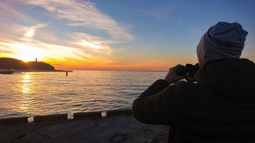 Rear view of man photographing sea against sky during sunset