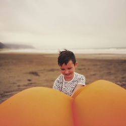 Boy with balloon sitting at beach against cloudy sky