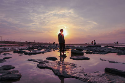 Silhouette of young man standing at beach against sky during sunset