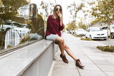 Portrait of young woman sitting on sidewalk by glass building