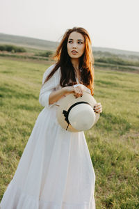 Thoughtful beautiful woman with sun hat standing on grassy land