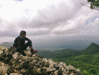 Full length of man looking at landscape while sitting on mountain against cloudy sky
