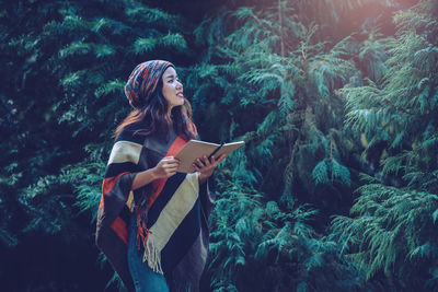 Side view of woman with book standing by trees in forest