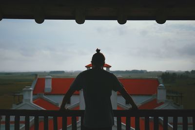 Rear view of mid adult man standing by railing in balcony against cloudy sky