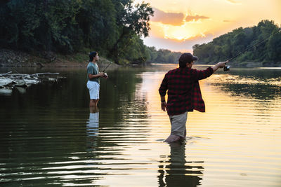 Male friends fishing while standing in lake during sunset