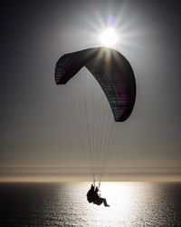 Silhouette people paragliding over sea against bright sky