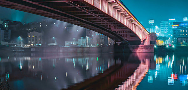 Illuminated bridge over river by buildings in city at night