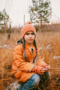 Pensive girl in orange coat sits in forest among dry grass and golden trees, outdoor, fall vibes