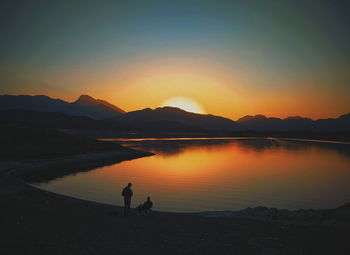 Silhouette people on lakeside against sky during sunset