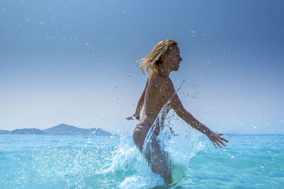 Side view of young woman splashing water in sea against sky