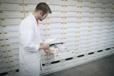 Side view of mature male pharmacist holding prescription paper standing by medicines in drawer at store