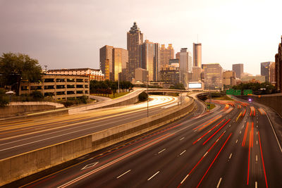 Light trails on road by modern buildings against sky in city