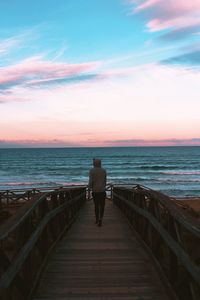 Rear view of man walking on boardwalk towards sea against sky during sunset