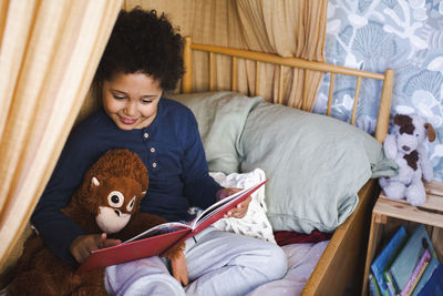 Boy reading story book while sitting with stuff toy on bed at home