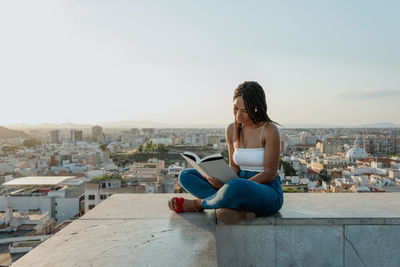 Trendy ethnic female in sandals reading textbook while sitting with crossed legs on fence in town