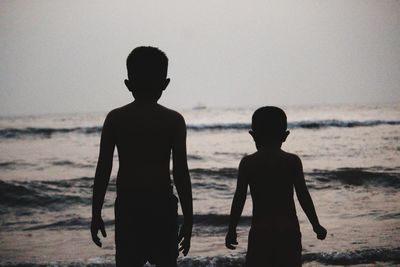 Rear view of silhouette boys standing at beach against clear sky