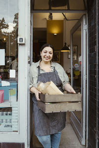 Portrait of happy female owner standing at doorway of deli holding crate