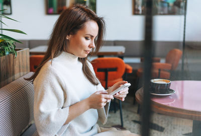 Attractive young brunette woman in white casual dress with cup of coffee using mobile phone in cafe