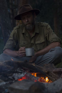 Man having drink while sitting by campfire during sunset