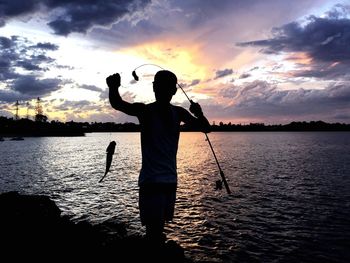 Silhouette fisherman with rod and fish by river against cloudy sky at dusk