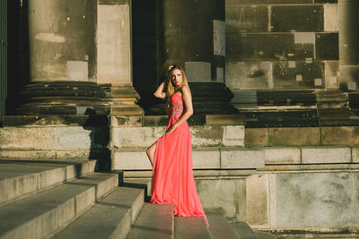 Full length side view of young woman in pink evening gown standing at on steps