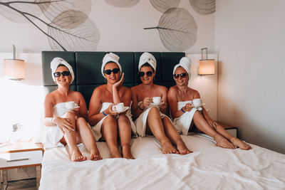 Four girls friends in white towels sitting on the bed having fun in hotel. vacation bloggers 