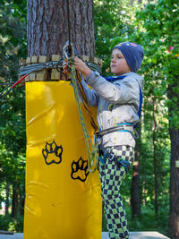 Full length of boy wearing safety harness while standing by tree trunk