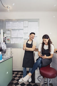 Female entrepreneur using smart phone while male colleague with arms crossed standing by in cafe