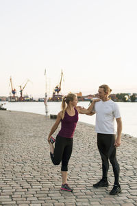 Man assisting woman in stretching on pier against clear sky