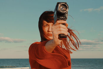 Woman looking away while photographing with camera against sea and sky